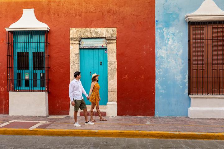 A couple walks past red and blue facades in the historic center of Campeche.