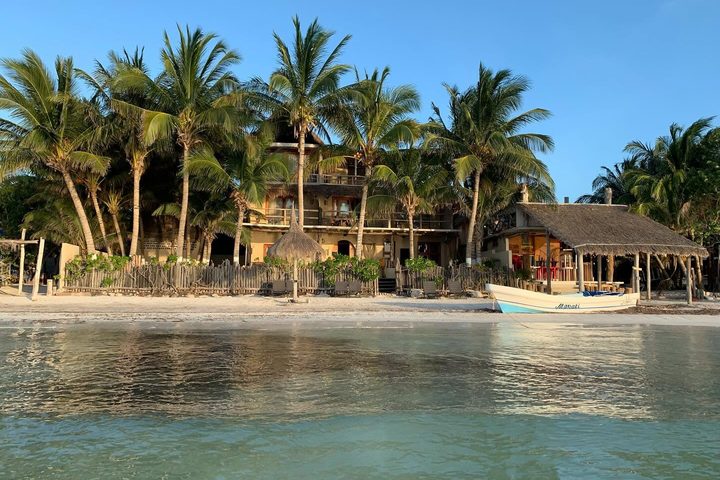 Casa Cat Ba vue depuis l’eau : la maison parmi les palmiers face à la plage de Holbox, avec un bateau sur le rivage.