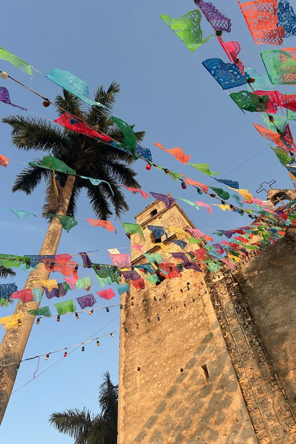 Papel picado de colores frente a la torre de la iglesia de Espita, junto a una palmera.